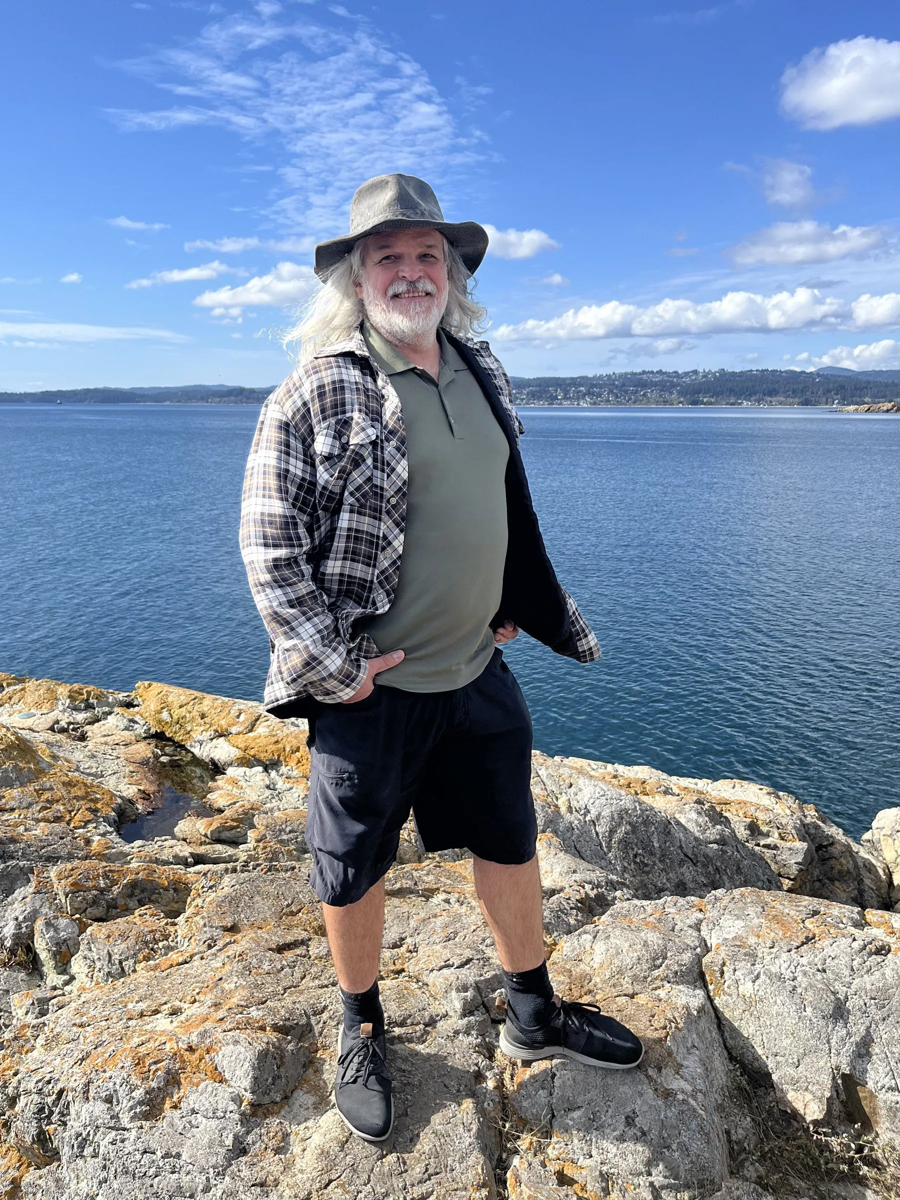 Greg on a rocky shore, in a hat and an open plaid shirt, with open water and a distant treeline behind him